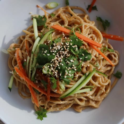 A vibrant bowl of cold sesame noodles, topped with colorful carrots, cucumbers, and fresh cilantro.