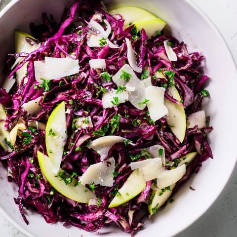 A rustic white bowl filled with colorful Red Cabbage Coleslaw with Apple and Parmesan, topped with shaved cheese and fresh parsley.